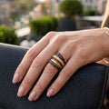A close-up of a hand wearing three stacked rings, resting on a black textured leather clutch with a blurred city background.